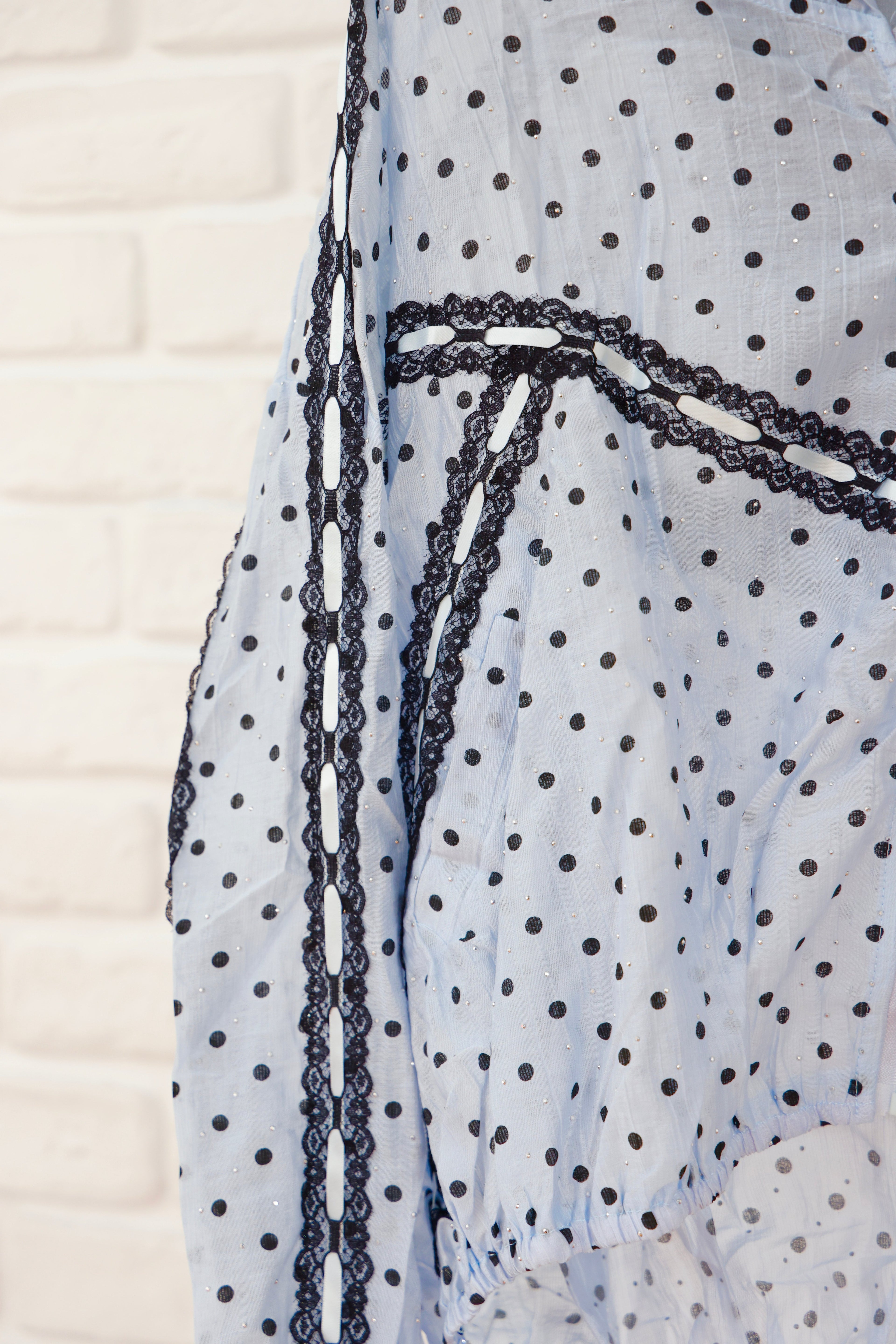 A close-up of a light blue sheer fabric with black polka dots and black lace trim, shown against a white brick wall background.