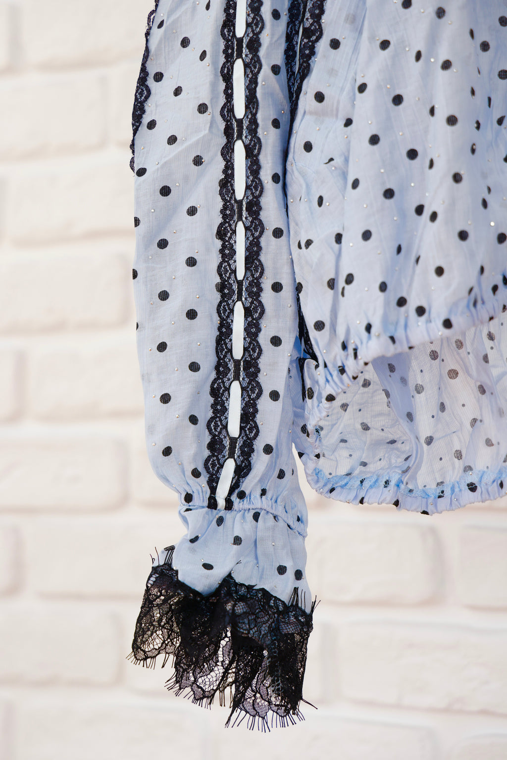 A close-up of a light blue sheer blouse with black polka dots, featuring black lace trim down the front and at the ruffled cuffs, set against a white brick wall background.