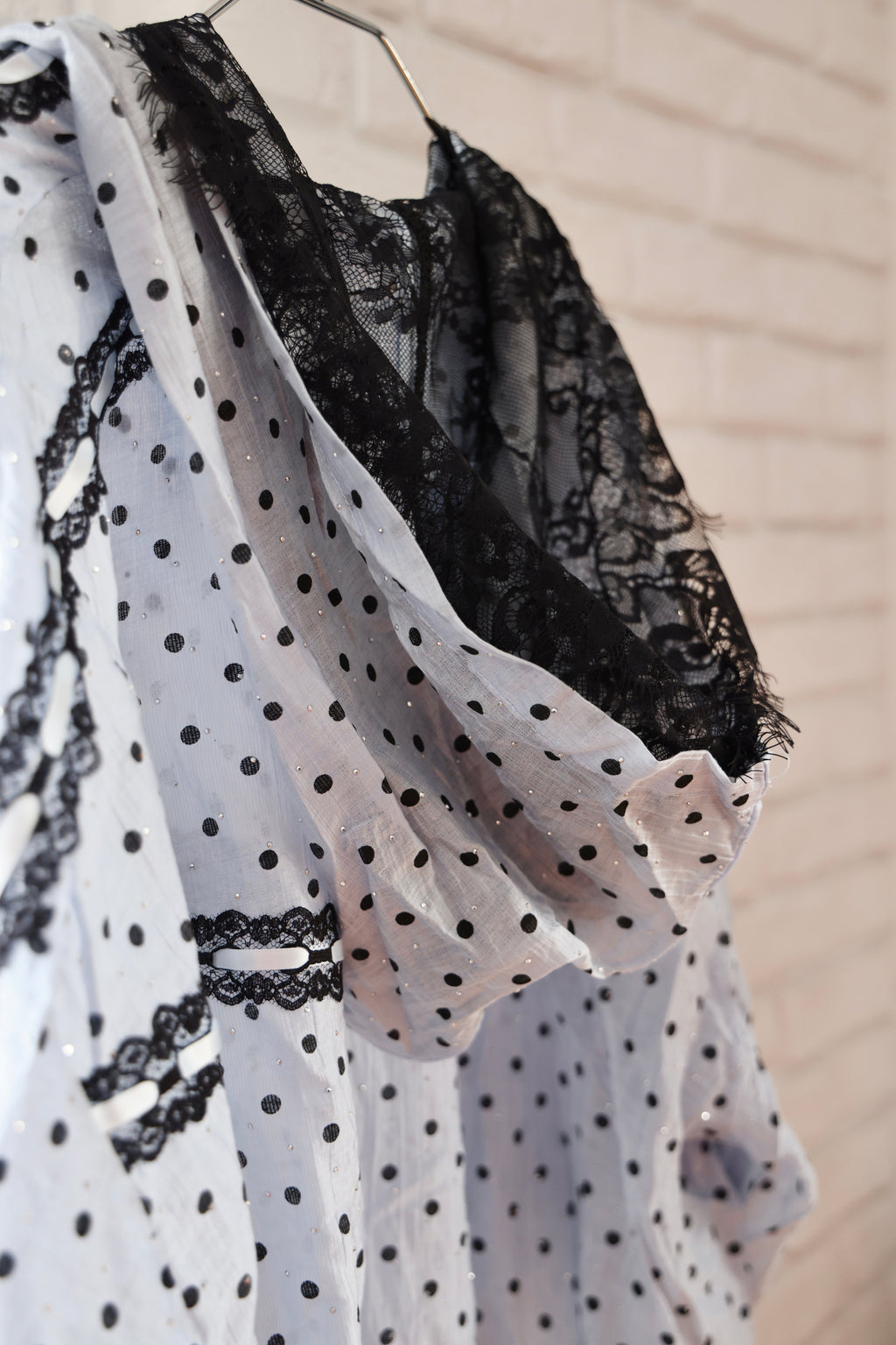 A sheer white polka-dot robe with black lace trim and a black lace shawl draped over it, hanging against a light brick wall.