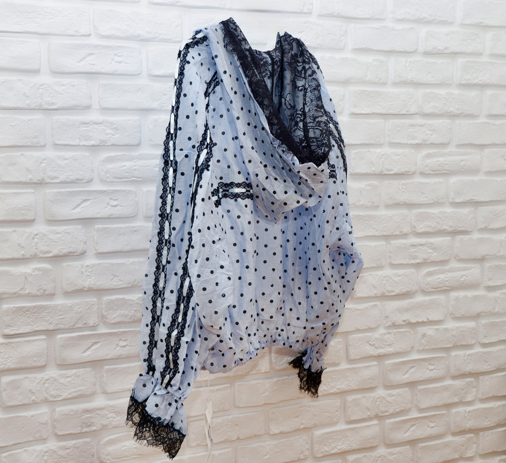 A sheer light blue and white polka dot scarf with black lace trim, draped over a white brick wall.