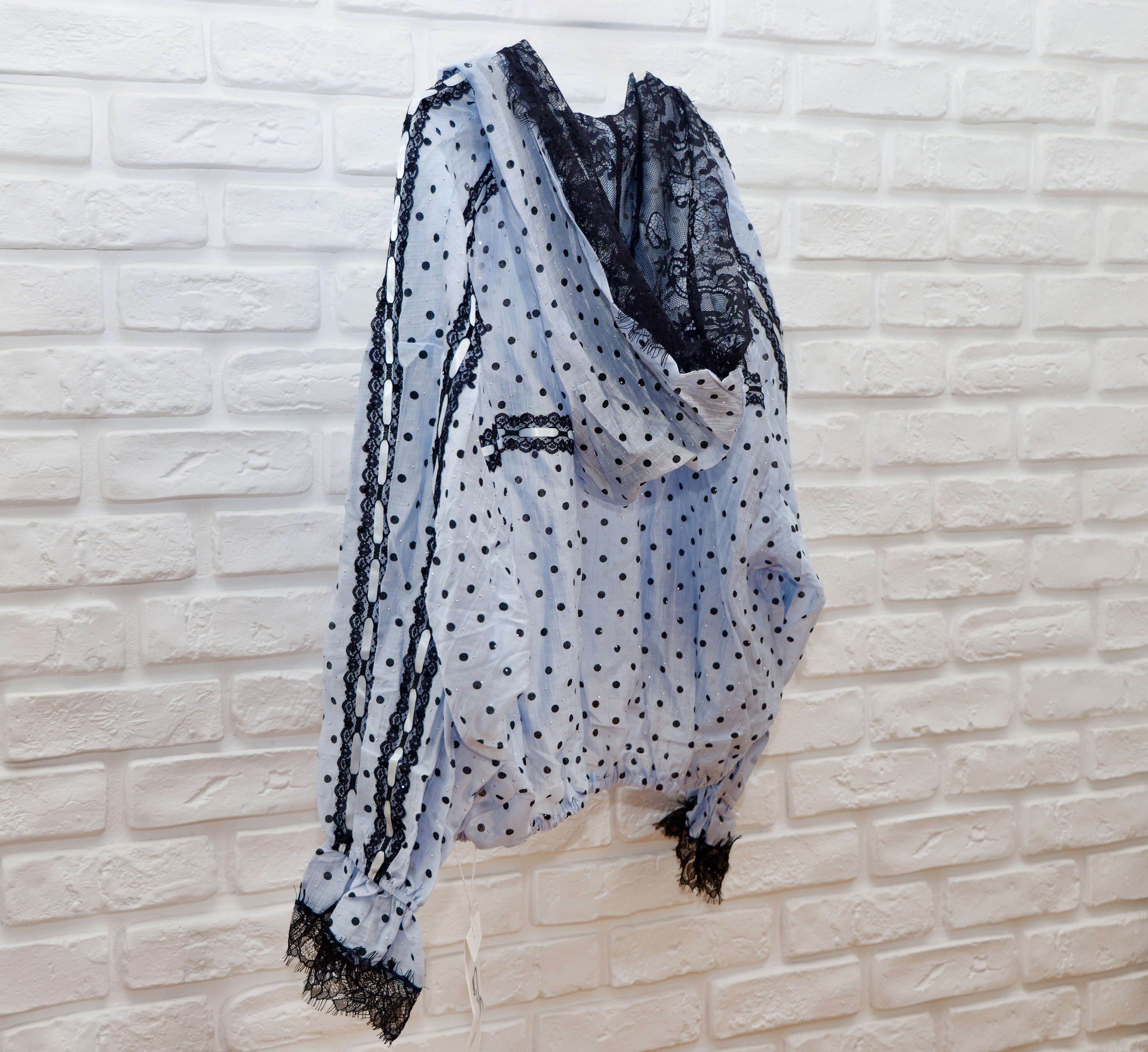 A sheer light blue and white polka dot scarf with black lace trim, draped over a white brick wall.