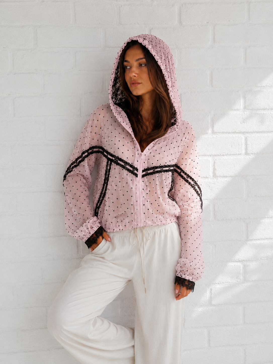 A woman wearing a light pink hooded jacket with black polka dots and black trim, paired with white drawstring sweatpants, posing against a white brick wall.