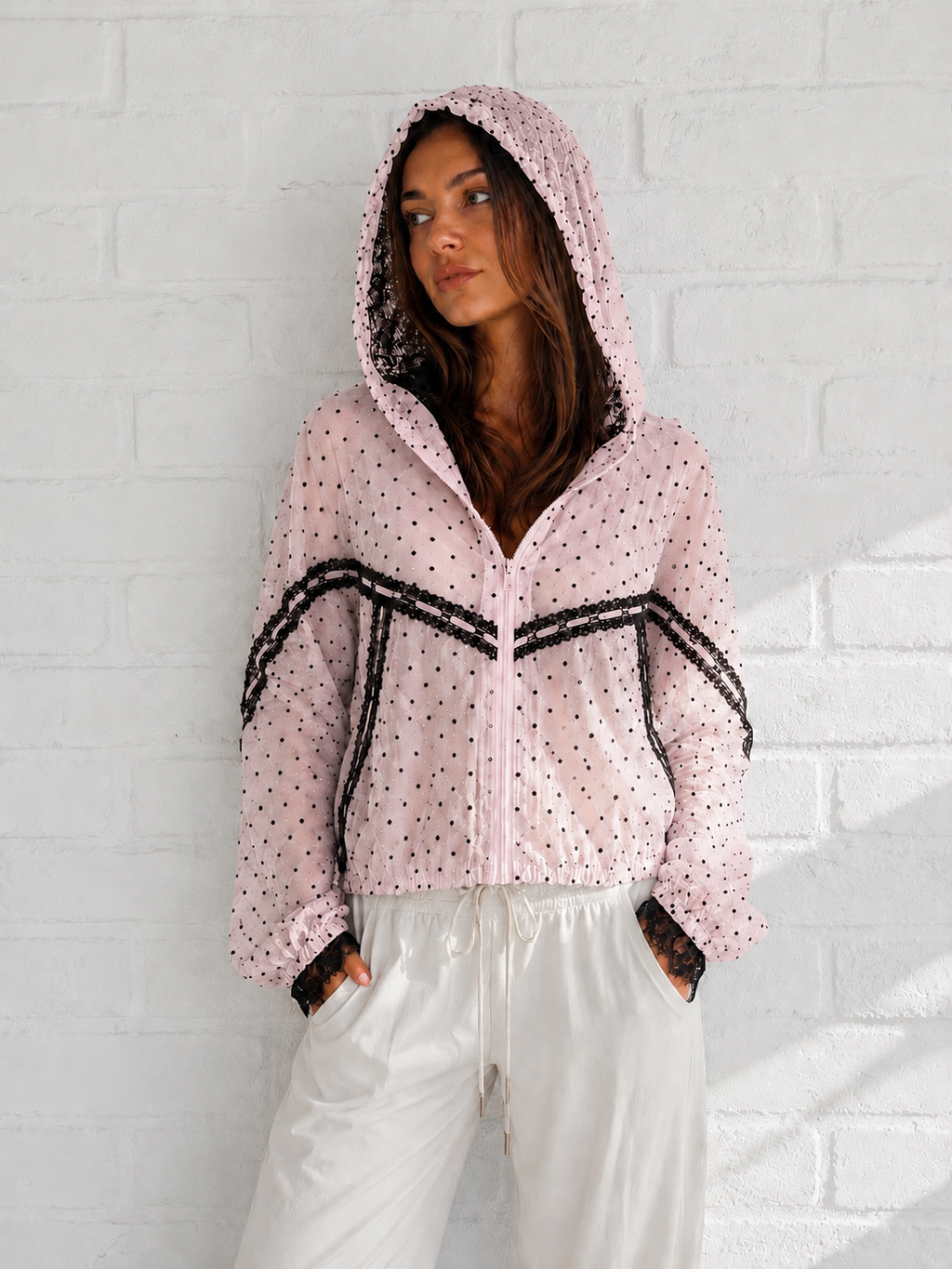 A woman wearing a light pink hooded zip-up jacket with black polka dots and black lace trim, paired with white pants, standing against a white brick wall.