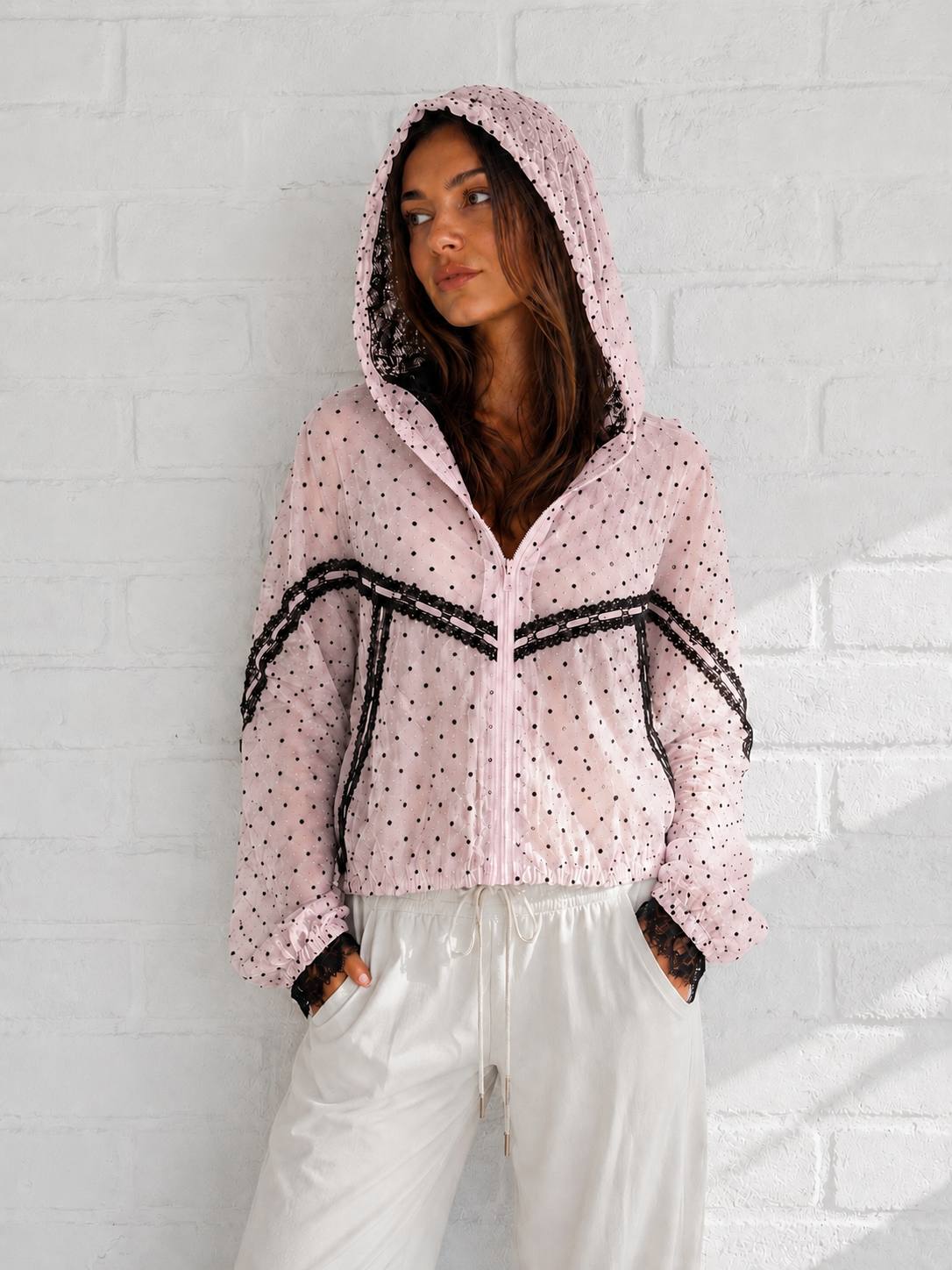 A woman wearing a light pink hooded zip-up jacket with black polka dots and black lace trim, paired with white pants, standing against a white brick wall.
