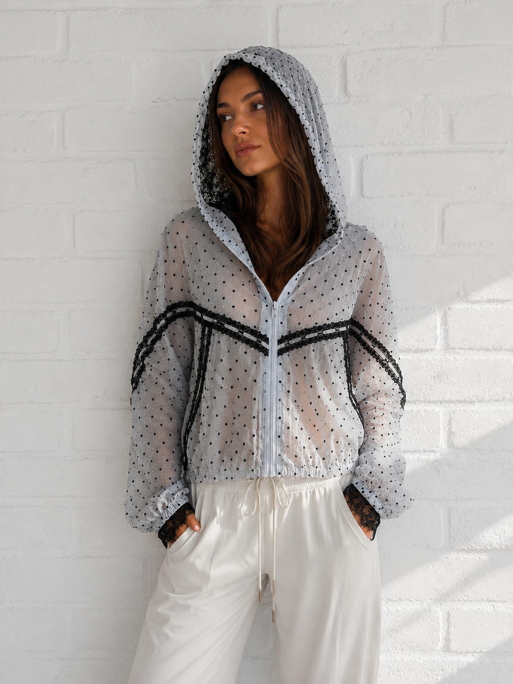A woman wearing a sheer white hooded jacket with black polka dots and black lace trim, paired with white drawstring pants, standing against a white brick wall.