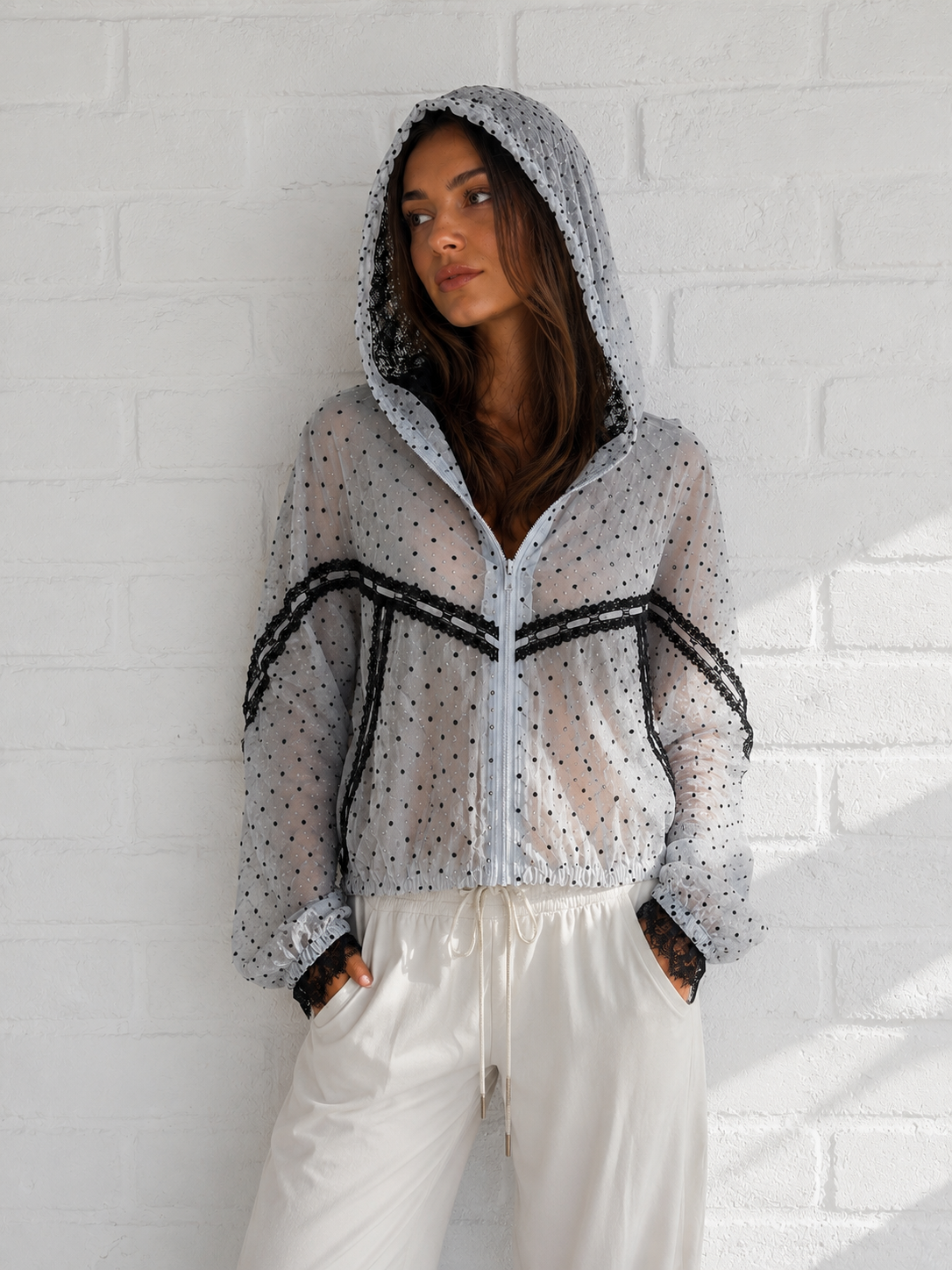 A woman wearing a sheer white hooded jacket with black polka dots and black lace trim, paired with white drawstring pants, standing against a white brick wall.