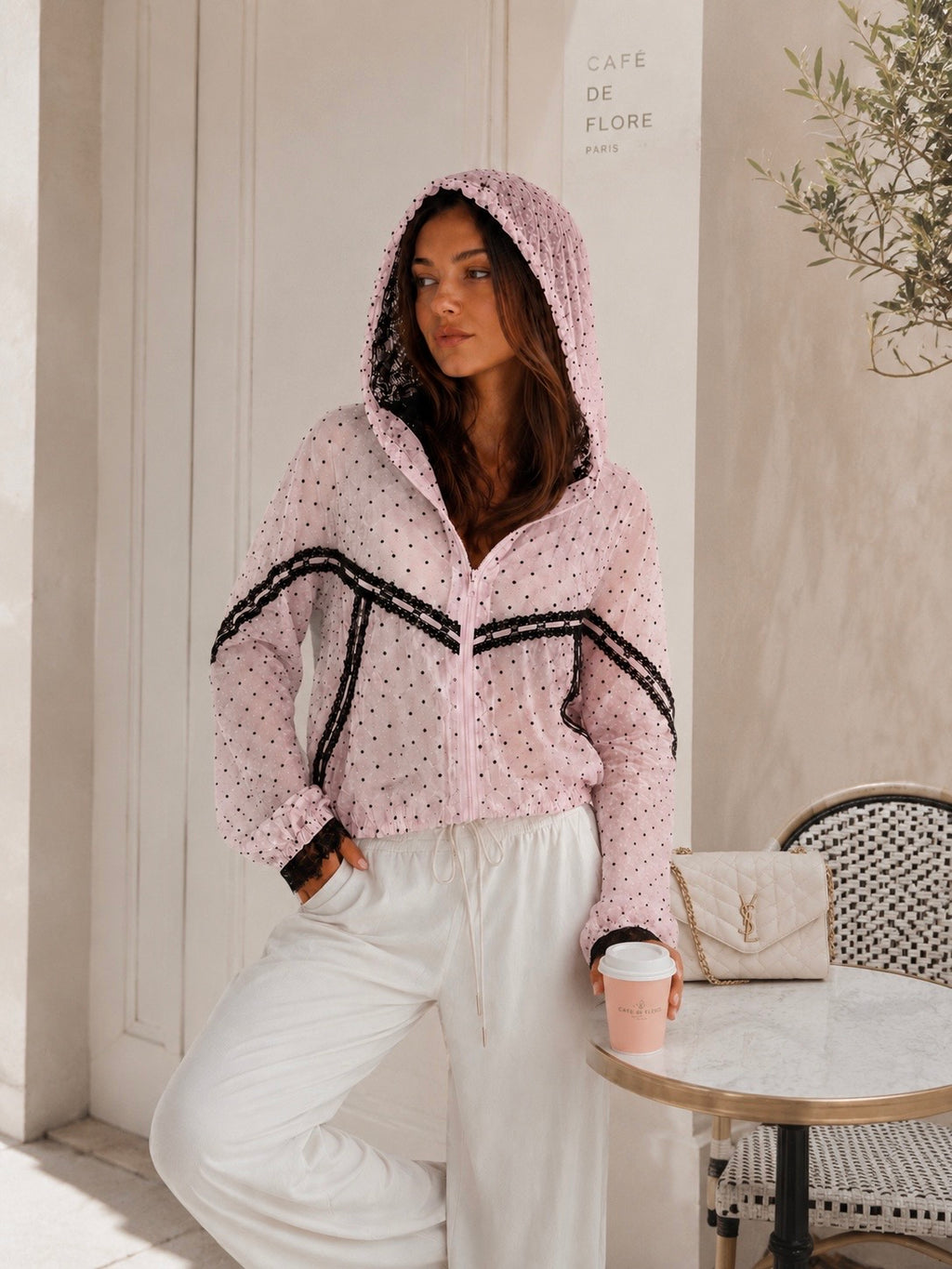 A woman models a light pink hooded zip-up jacket with black trim and white polka dots, paired with white sweatpants, in a bright, modern indoor setting.
