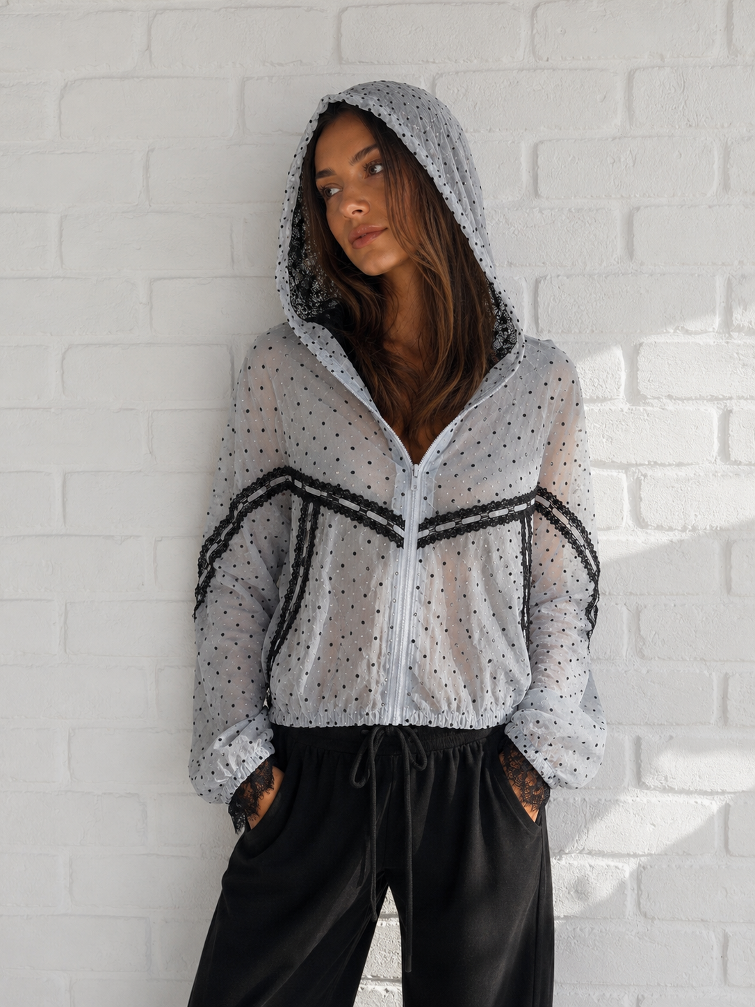 A woman wearing a sheer light gray zip-up hoodie with black polka dots and black stripe accents, paired with black sweatpants, posing against a white brick wall.