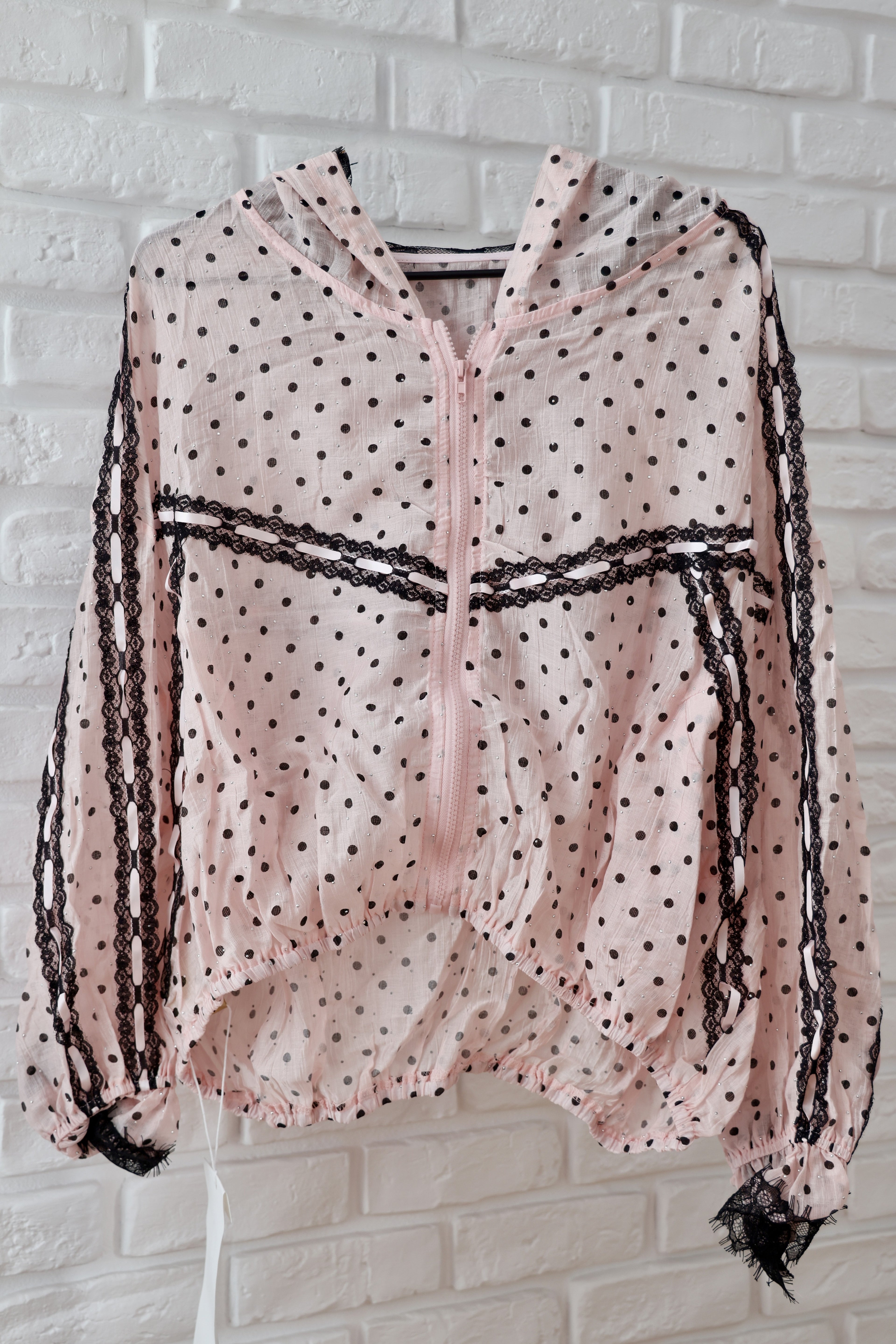 A light pink hooded jacket with black polka dots, black lace trim along the zipper and sleeves, and a white brick wall background.