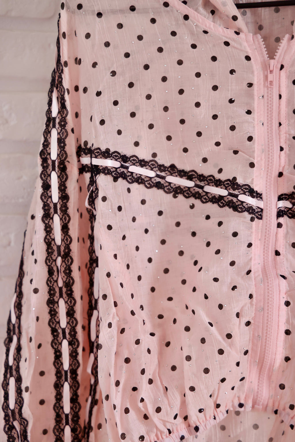 A close-up of a light pink sheer fabric with black polka dots, featuring black lace trim and a pink zipper.