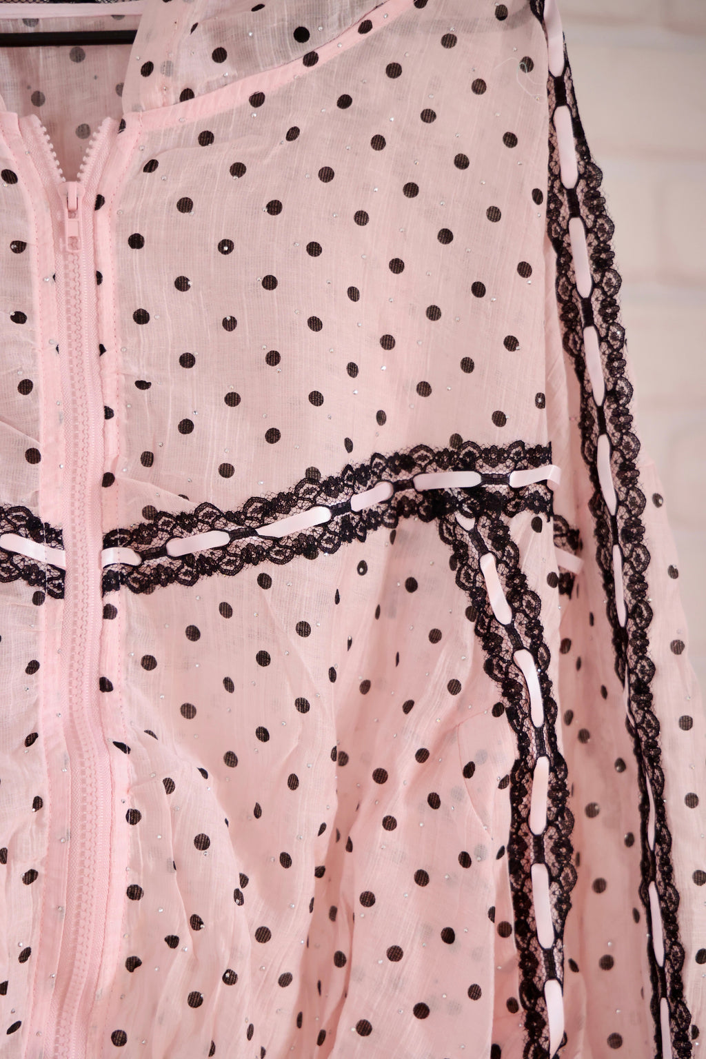 A close-up of a light pink sheer mesh garment with black polka dots and black lace trim along a zipper and side edges.