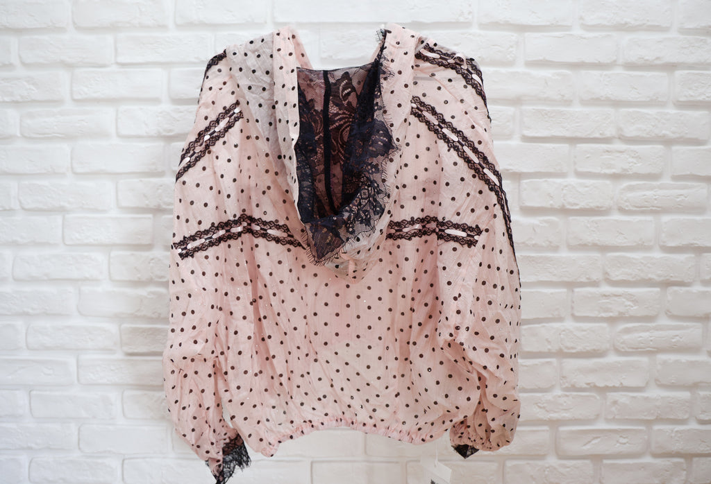 A light pink sheer blouse with black polka dots, black lace trim, and a black underlayer, displayed against a white brick wall.